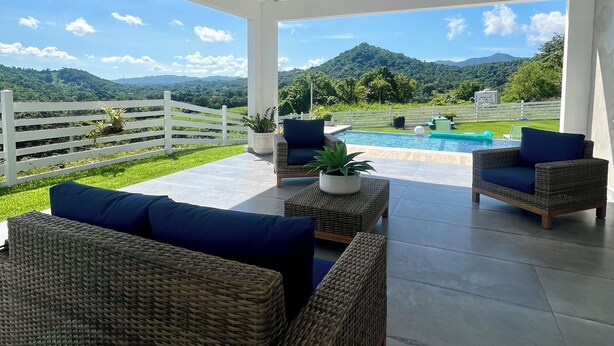 Un espacio al aire libre techado con mobiliario de mimbre, un ventilador de techo y vistas a una piscina y montañas.
