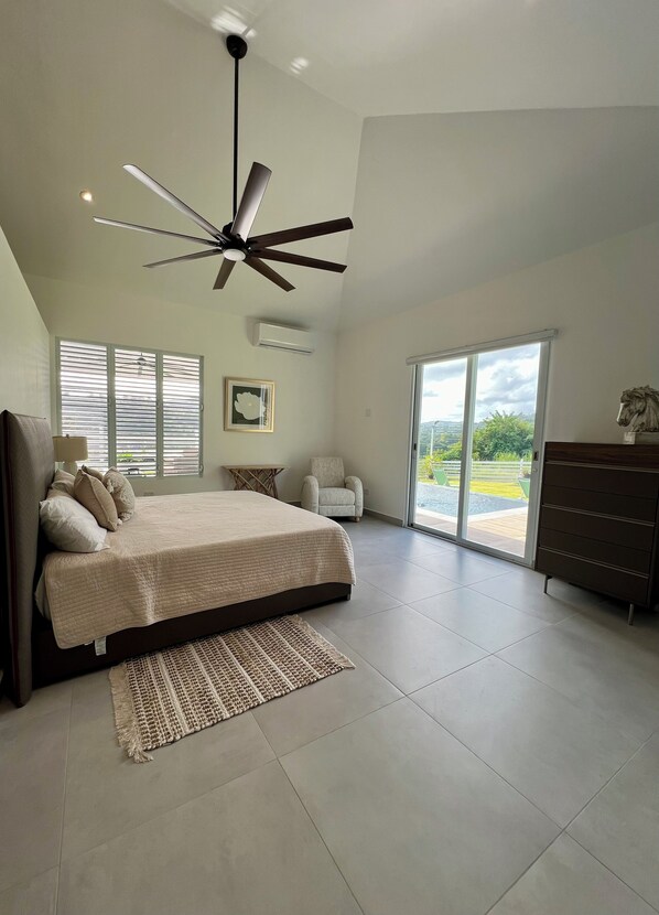A modern bedroom with a large bed, a ceiling fan, a view through the glass door, and a dresser.
