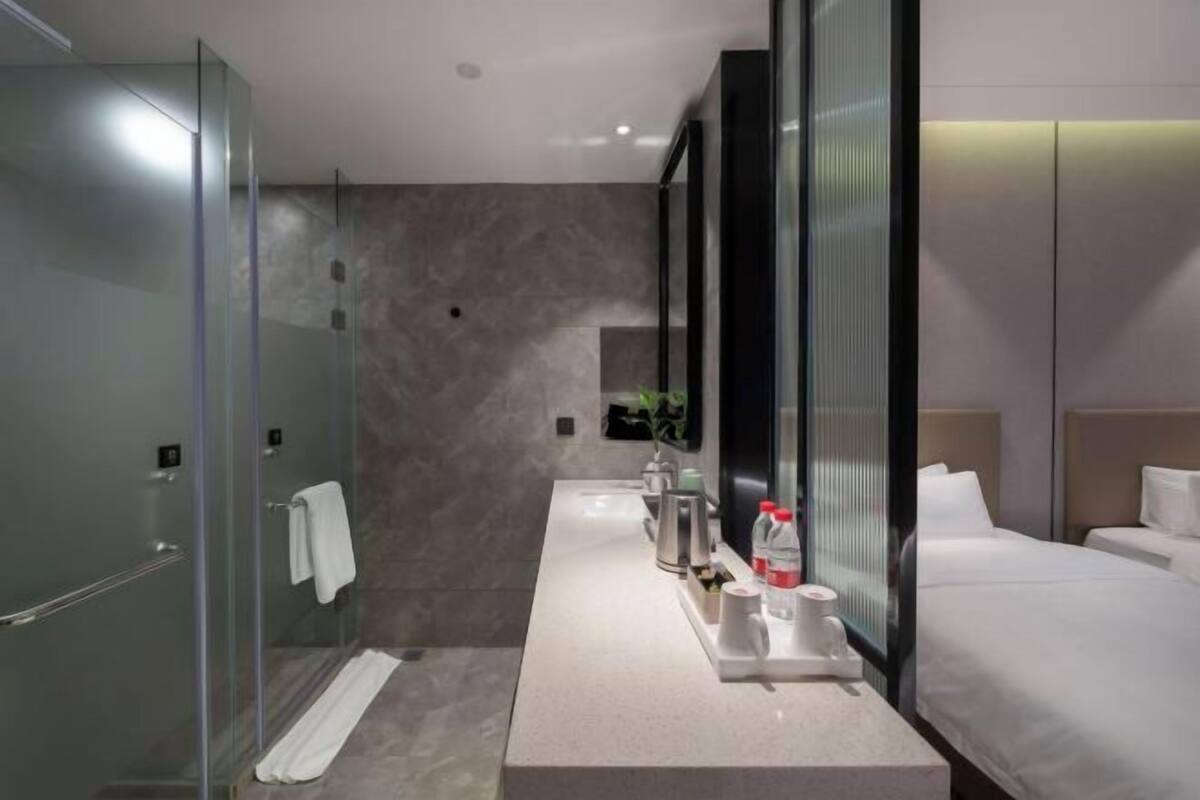 Een moderne hotelbadkamer met een douchecabine van glas, een wastafel met toiletartikelen, en een bed met witte lakens.