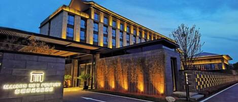 Un hotel moderno con una entrada amplia, una fachada bien iluminada y una zona ajardinada con árboles.