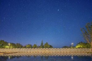 Een sterrenhemel boven een spiegelbeeldig waterbassin, omgeven door bomen en een bakstenen muur.