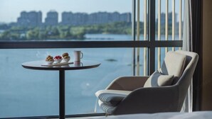 A modern room with a large window overlooking a body of water and buildings. There is a round table with a cup and a bowl of fruit. - Wuhu Maison New Century Hotel (Wuhu)
