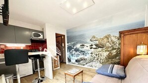 A modern living room with a large ocean view mural, a sofa, and a small wooden table. - Studio archi cosy – 5min des plages (Quiberon)