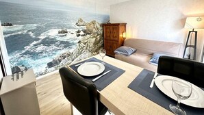 A cozy room with a view of the ocean, a dining table set with plates and cutlery, a sofa, and a cabinet. - Studio archi cosy – 5min des plages (Quiberon)