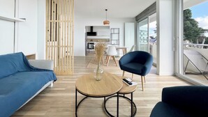 Ein modernes Wohnzimmer mit einem blauen Sofa, einem runden Holztisch und einem blauen Sessel. Im Hintergrund eine Holzvertäfelung und durch große Fenster der Blick auf einen Balkon.