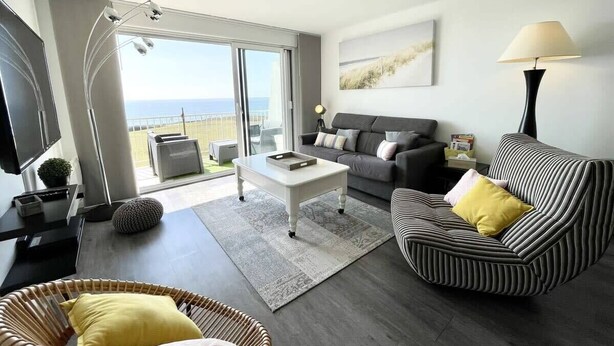 Ein modernes Wohnzimmer mit Sofa, Sessel und Couchtisch. Ein großes Fenster bietet Blick ins Freie.