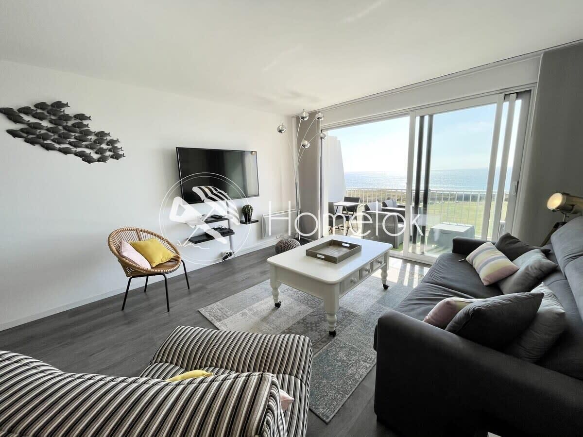 Ein modernes Wohnzimmer mit Sofa, Sessel und Couchtisch. Ein Flachbildfernseher ist an der Wand montiert, und ein großes Fenster bietet Meerblick.