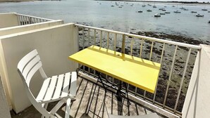 Un balcon avec une table jaune et des chaises blanches, donnant sur une mer calme où voguent des bateaux.