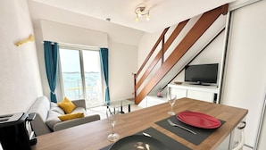 Une chambre d’hôtel moderne avec une table à manger en bois dressée pour deux personnes, un canapé avec des coussins jaunes, une télévision et une vue sur la mer par de grandes fenêtres.