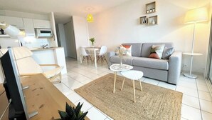 Un salon moderne avec un canapé gris, une petite table blanche et une table basse. On y trouve également un coin cuisine avec des meubles blancs et un four à micro-ondes. Une suspension jaune est suspendue au-dessus de la table à manger.
