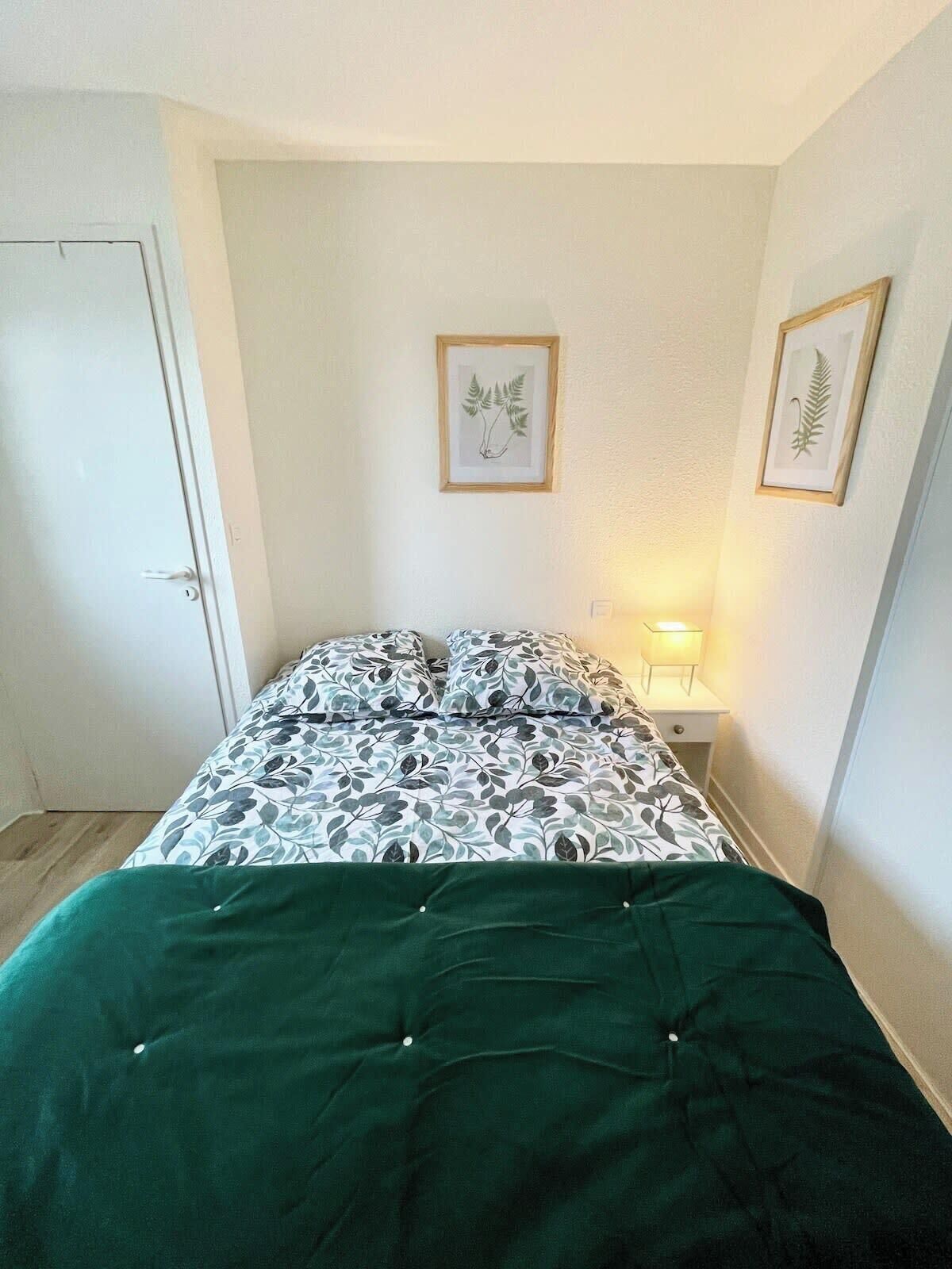 A bedroom with a bed, a nightstand, a door, and two framed botanical prints on the wall.
