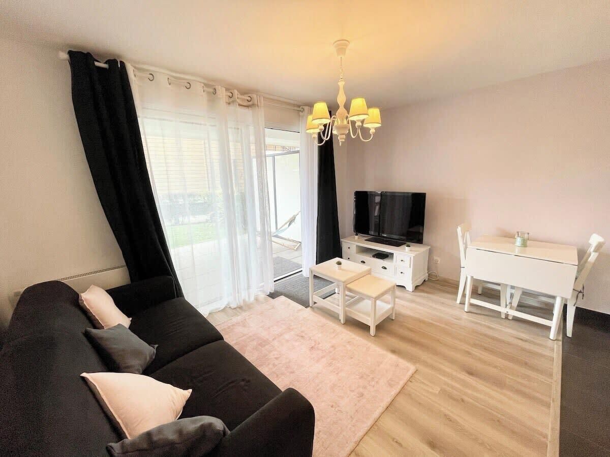 A modern living room with a black sofa, a small white table, a TV, and a dining area with a white table and chairs.