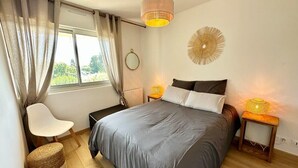 A bedroom with a large bed, a bedside table with a lamp, a chair, a round mirror, and a window with curtains.