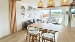 Eine moderne Küche mit einem Holztisch und Barhocker, ein Essbereich mit einer Couch und Stühlen sowie ein Blick ins Freie durch große Fenster.
