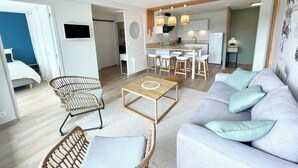 Ein modernes Wohnzimmer mit einer Couch, zwei Korbstühlen, einem Couchtisch und einem Essbereich, im Hintergrund ist eine Küche zu sehen.