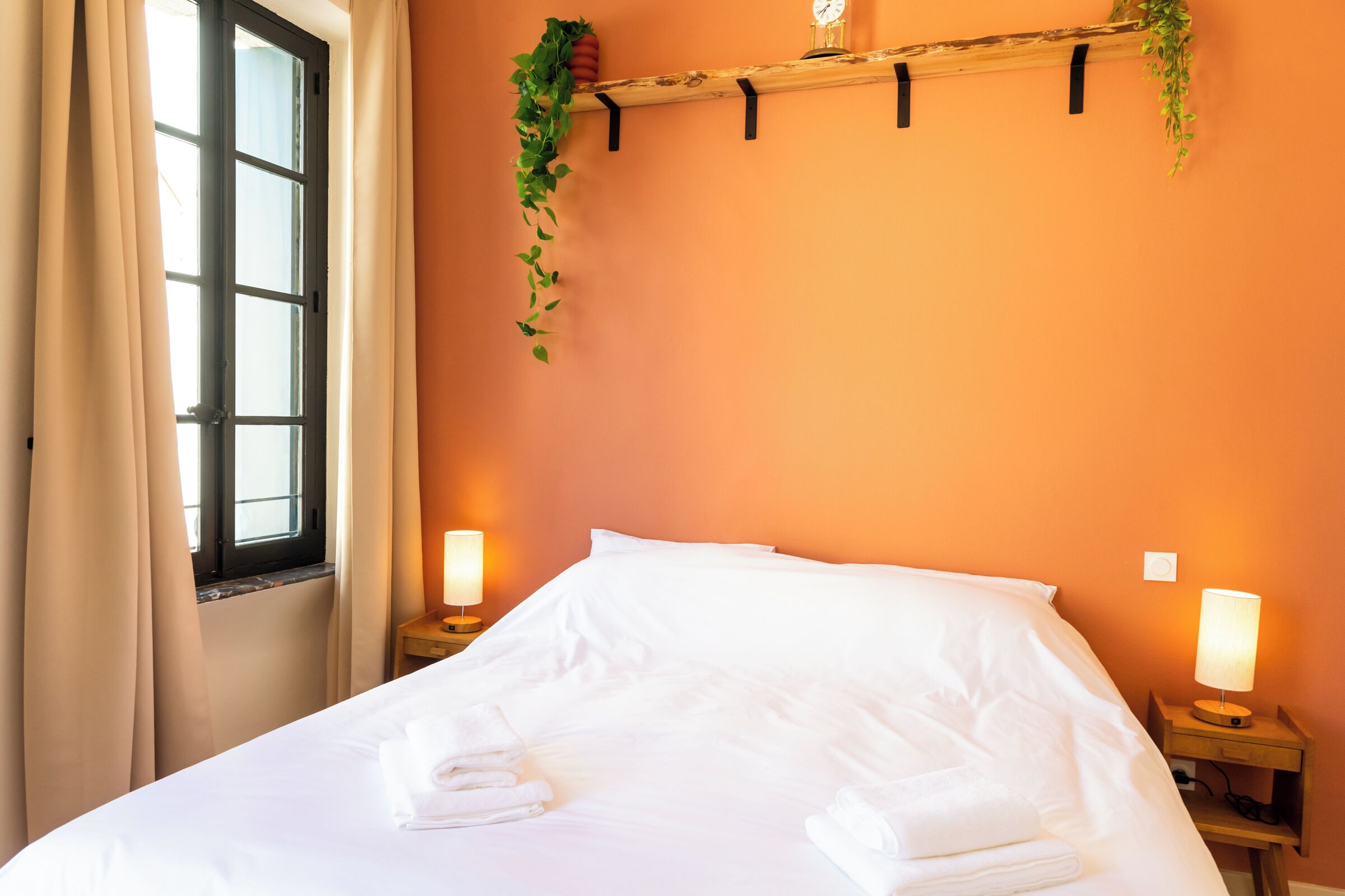 Cama bem arrumada com lençóis e toalhas brancos, duas luminárias de cabeceira, uma janela com cortinas e uma prateleira fixa na parede com plantas.