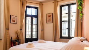 A hotel room with a large bed, two windows with curtains, a bedside table with books, and a wall-mounted plant.