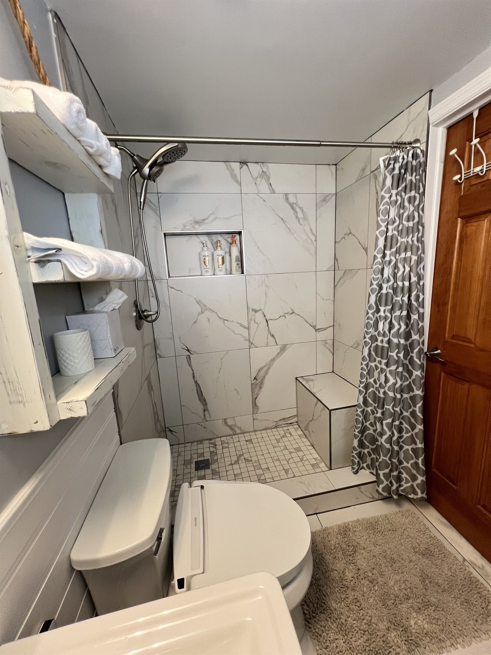 A bathroom with a toilet, a shower, and a sink. There are shelves with toiletries and a towel rack.