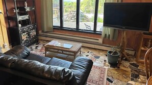 A living room with a leather couch, a television, a large window with a view of trees, and a decorative cabinet. - Magic Forest Farm's Mountain View Suite (Coeymans Hollow)