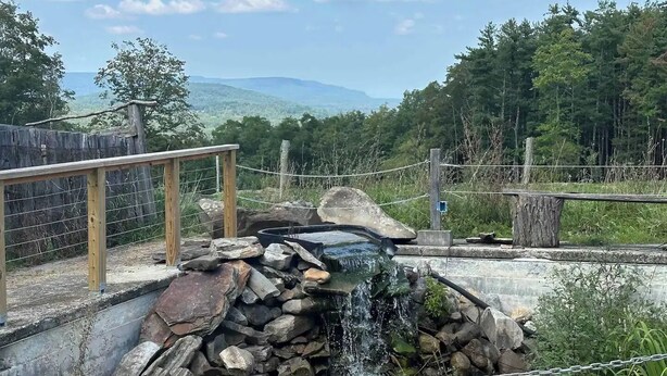 A natural water feature with a wooden deck and chain-link fence, surrounded by trees and mountains. - Magic Forest Farm's Mountain View Suite (Coeymans Hollow)
