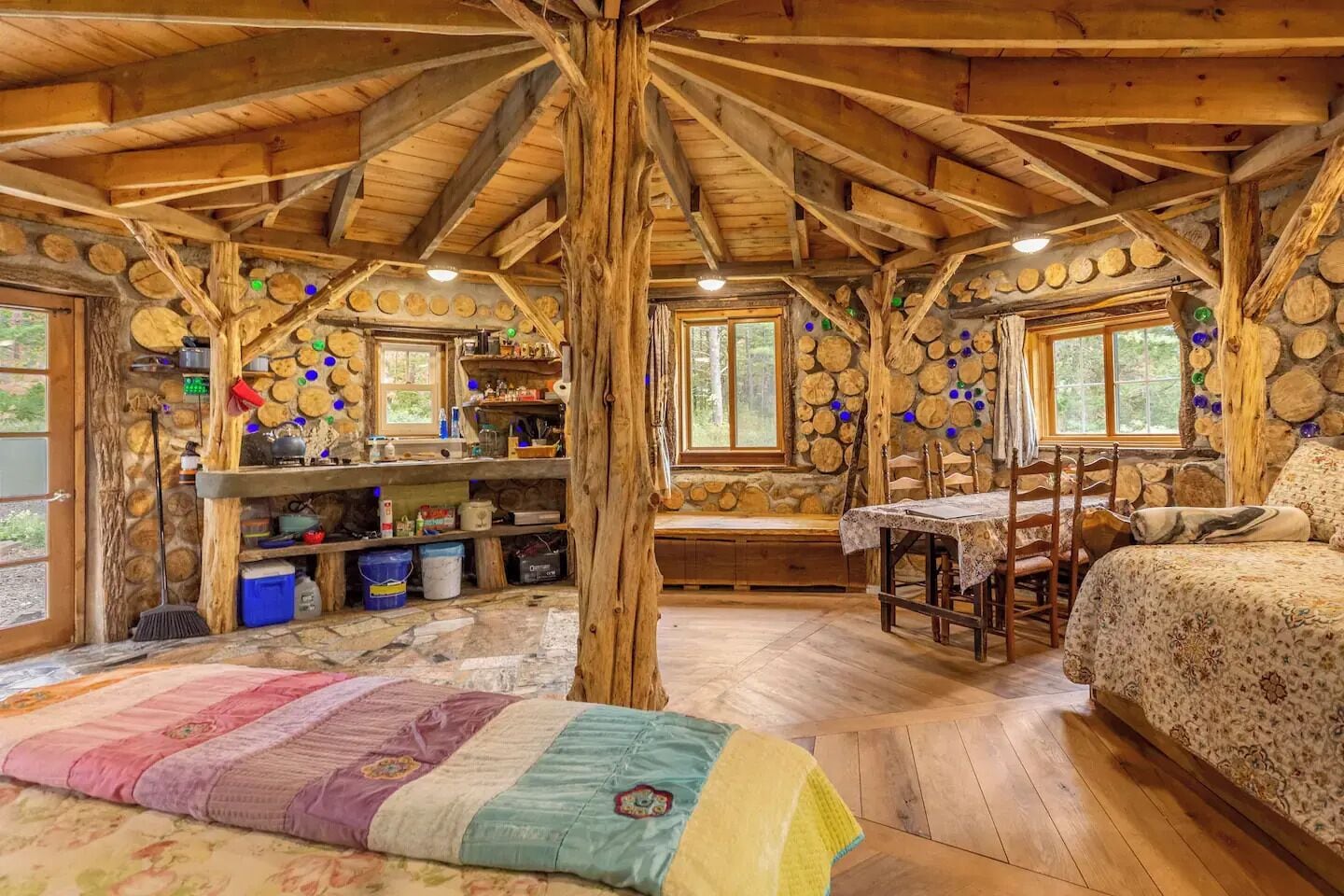 Interior rústico de una cabaña con techo de madera, paredes de piedra, un banco de madera, una mesa y una cama.
