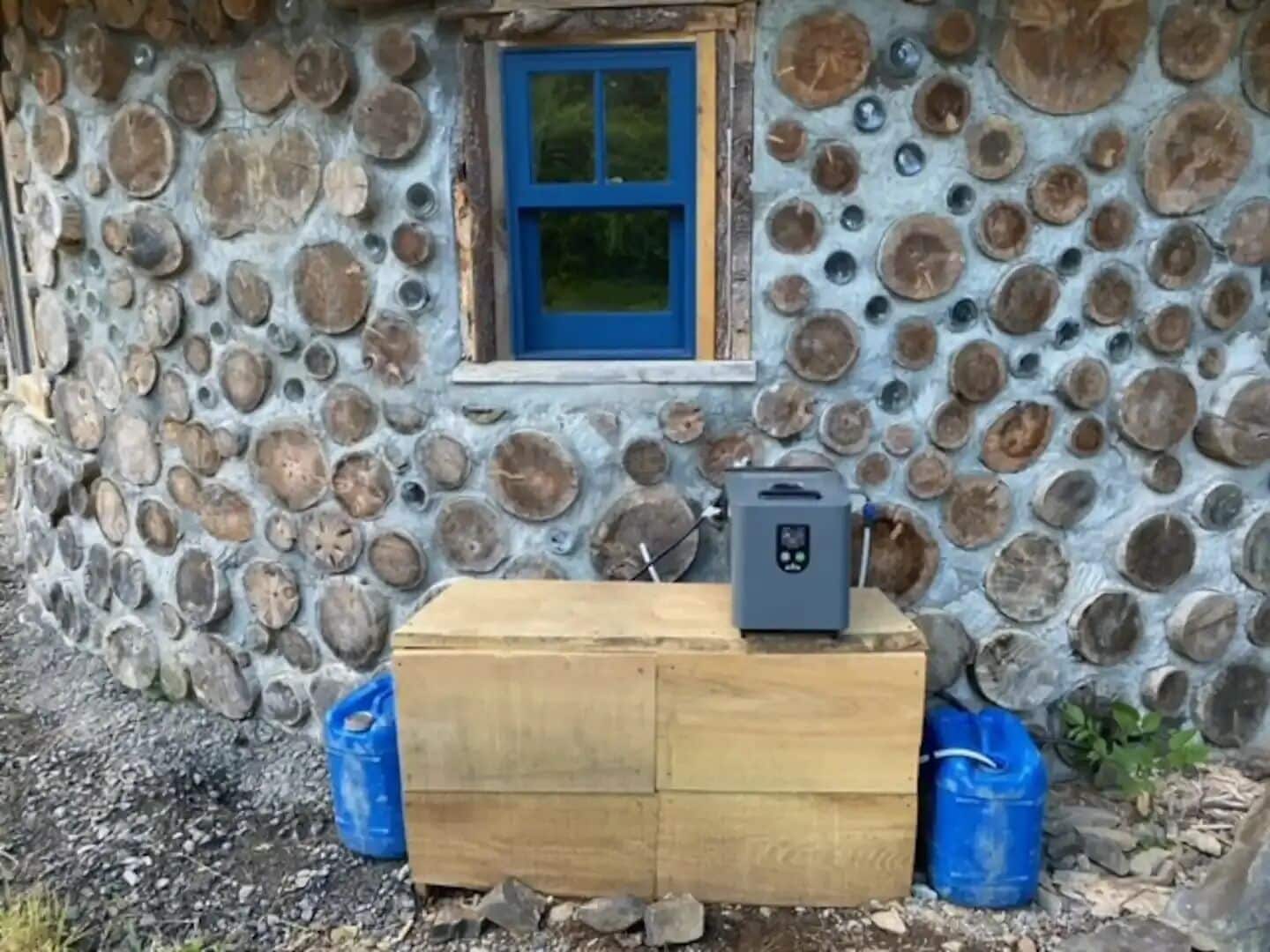 Un rustico chalet con facciata in pietra, una piccola finestra con cornice blu e un termosifone portatile su un baule di legno.