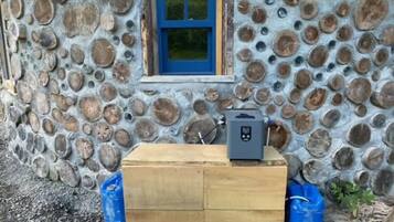 Un rustico chalet con facciata in pietra, una piccola finestra con cornice blu e un termosifone portatile su un baule di legno.