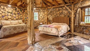 Ein rustikales Blockhaus-Interieur mit Steinwand, Holzbalken, einem Bett, einer Couch und einem großen Baumstumpf in der Mitte.