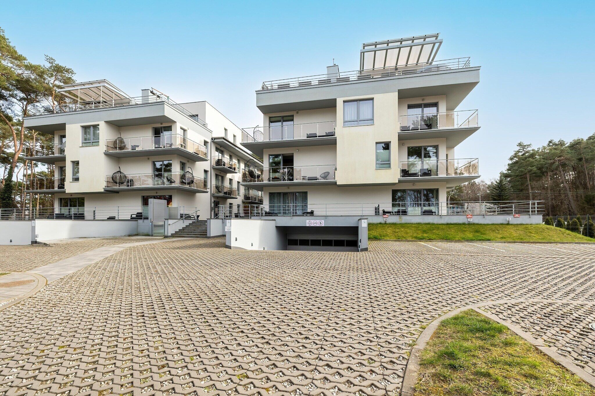 A modern multi-story residential building with balconies, a paved courtyard, and a clear sky.