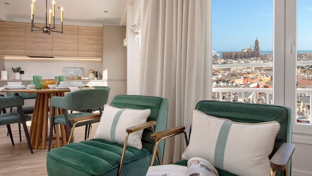 Un salon moderne avec vue sur le paysage urbain, comprenant des fauteuils recouverts de tissu vert, une table à manger en bois et un lustre.
