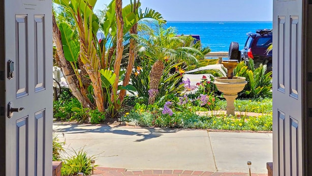 A view through open doors to a garden with tropical plants, a fountain, and a parking area with a vehicle. - Ocean Front+ Theater Room +Hot Tub + Game Room (Carlsbad)