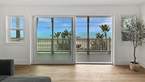 A room with a wooden floor, a grey sofa, a balcony with a view of the ocean, and a potted plant. - Ocean Front+ Theater Room +Hot Tub + Game Room (Carlsbad)