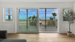 A room with a wooden floor, a grey sofa, a balcony with a view of the ocean, and a potted plant. - Oceanfront- Views from balcony-Movie Room-Hot tub (Carlsbad)