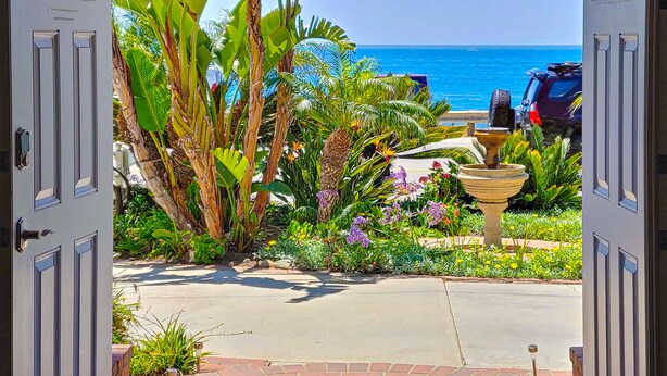 A view through open doors to a garden with tropical plants, a fountain, and a parking area with a vehicle. - Oceanfront- Views from balcony-Movie Room-Hot tub (Carlsbad)
