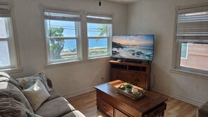 Ein Wohnzimmer mit Fernseher auf einem Holzboden, einer Couch und großen Fenstern mit Meerblick.