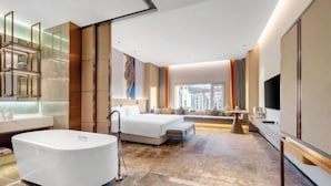 A modern hotel room with a large bed, a separate seating area, a bathtub, and a large window offering a city view.