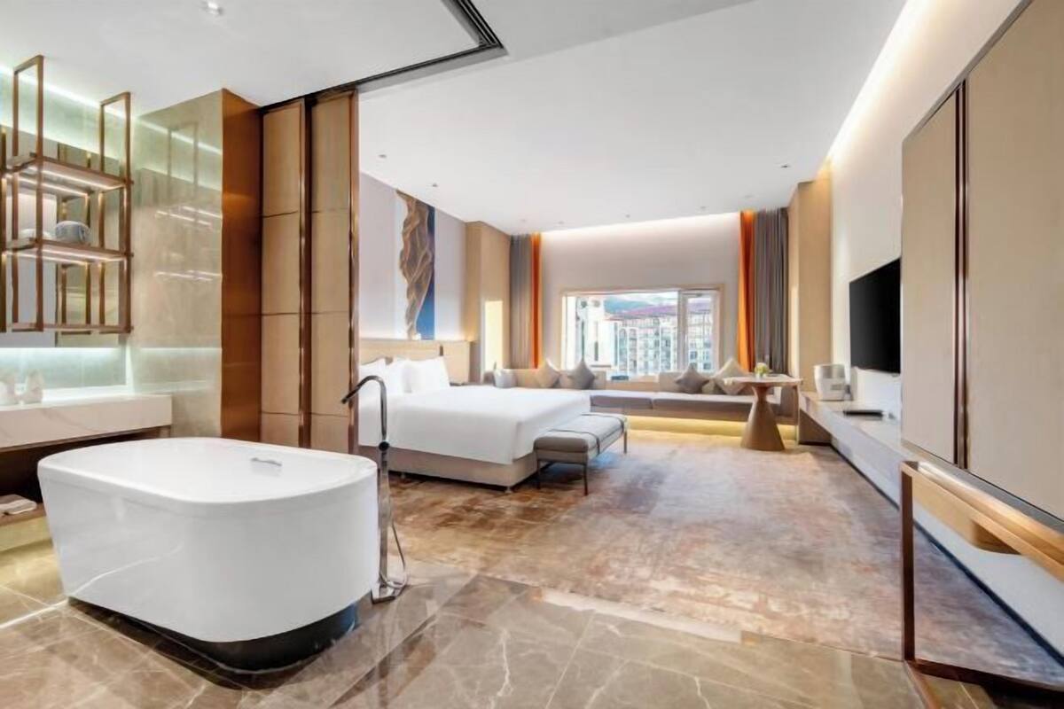 A modern hotel room with a large bed, a separate seating area, a bathtub, and a large window offering a city view.