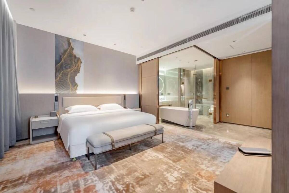 A modern hotel room with a large bed, bedside tables, a bench, a glass shower, and a walk-in closet.