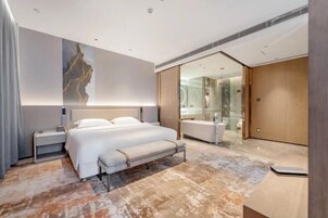 Una habitación de hotel moderna con una cama grande, mesitas de noche, un banco, ducha de vidrio y vestidor.
