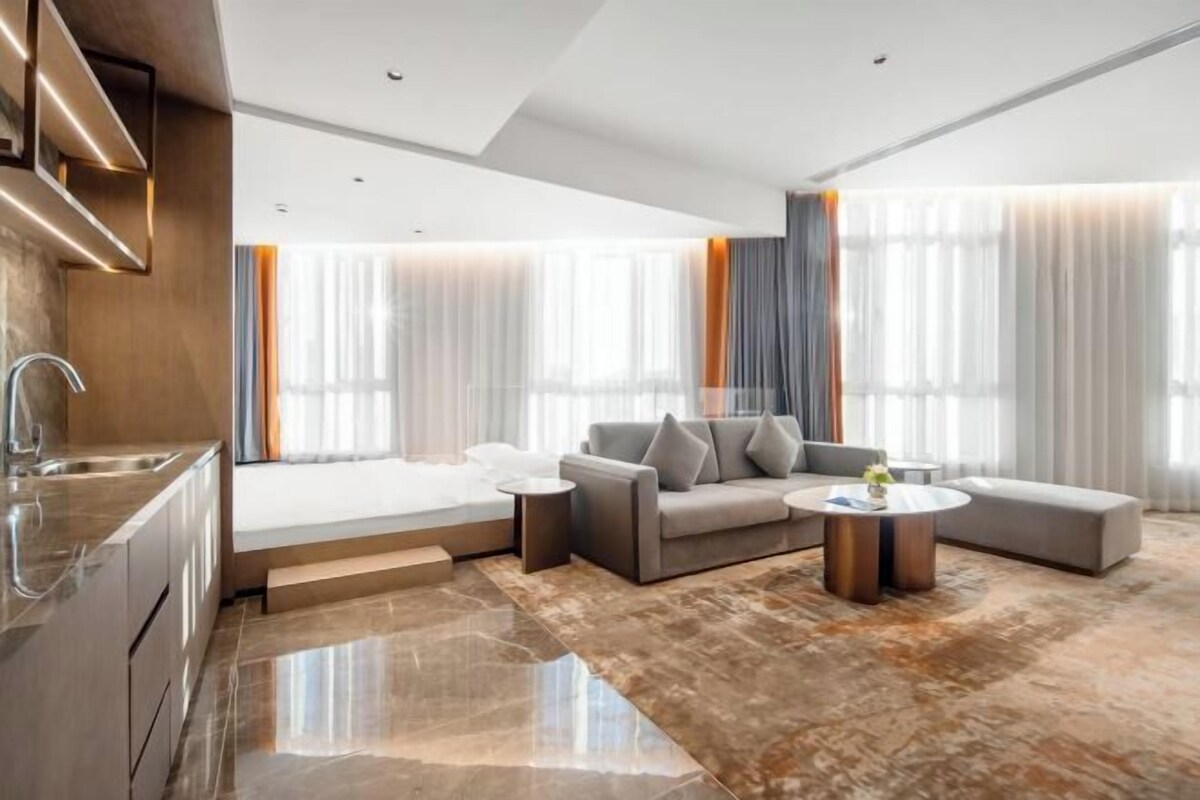 Quarto de hotel moderno com cozinha, sofá, mesa de centro e janelas amplas com cortinas.
