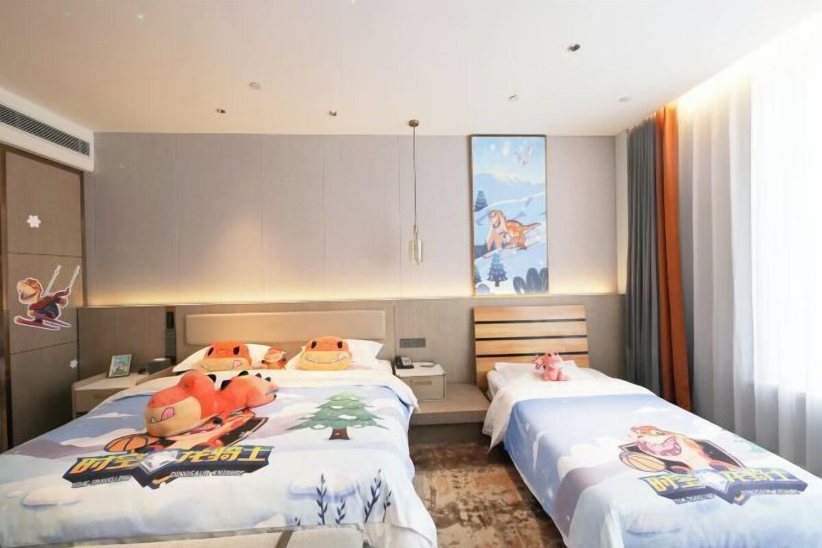 Quarto de hotel com duas camas, cada uma com colcha de tema de desenho animado. Há uma pintura grande na parede acima das camas, uma mesa de cabeceira com abajur e uma janela com cortinas.