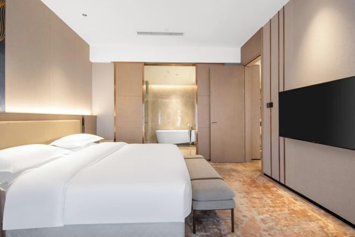 Quarto de hotel moderno com uma cama grande, um banco, uma TV de tela plana e um banheiro visível através de uma porta de vidro.