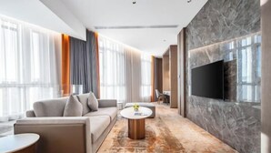 A modern hotel room with a sofa, a round coffee table, a flat-screen TV mounted on the wall, and a large window with curtains.