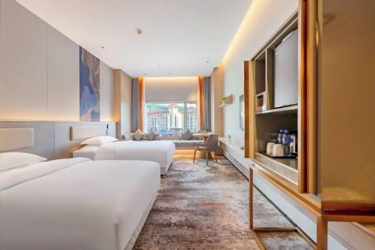 A hotel room with two beds, a desk, a chair, a TV, and a large window.