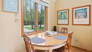 Un coin repas avec une table ronde, quatre chaises en bois, une fenêtre donnant sur les arbres et trois œuvres d’art encadrées accrochées au mur.