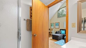 Une pièce avec une porte en bois, un mur blanc, un plafond doté d’une lucarne, un canapé noir, un tapis bleu et un lavabo blanc.