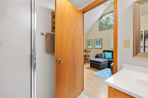 A room with a wooden door, a white wall, a ceiling with a skylight, a black sofa, a blue rug, and a white sink.