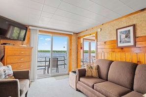 A room with a wooden interior, a television, a sofa, a chair, a fireplace, and a view of a lake.