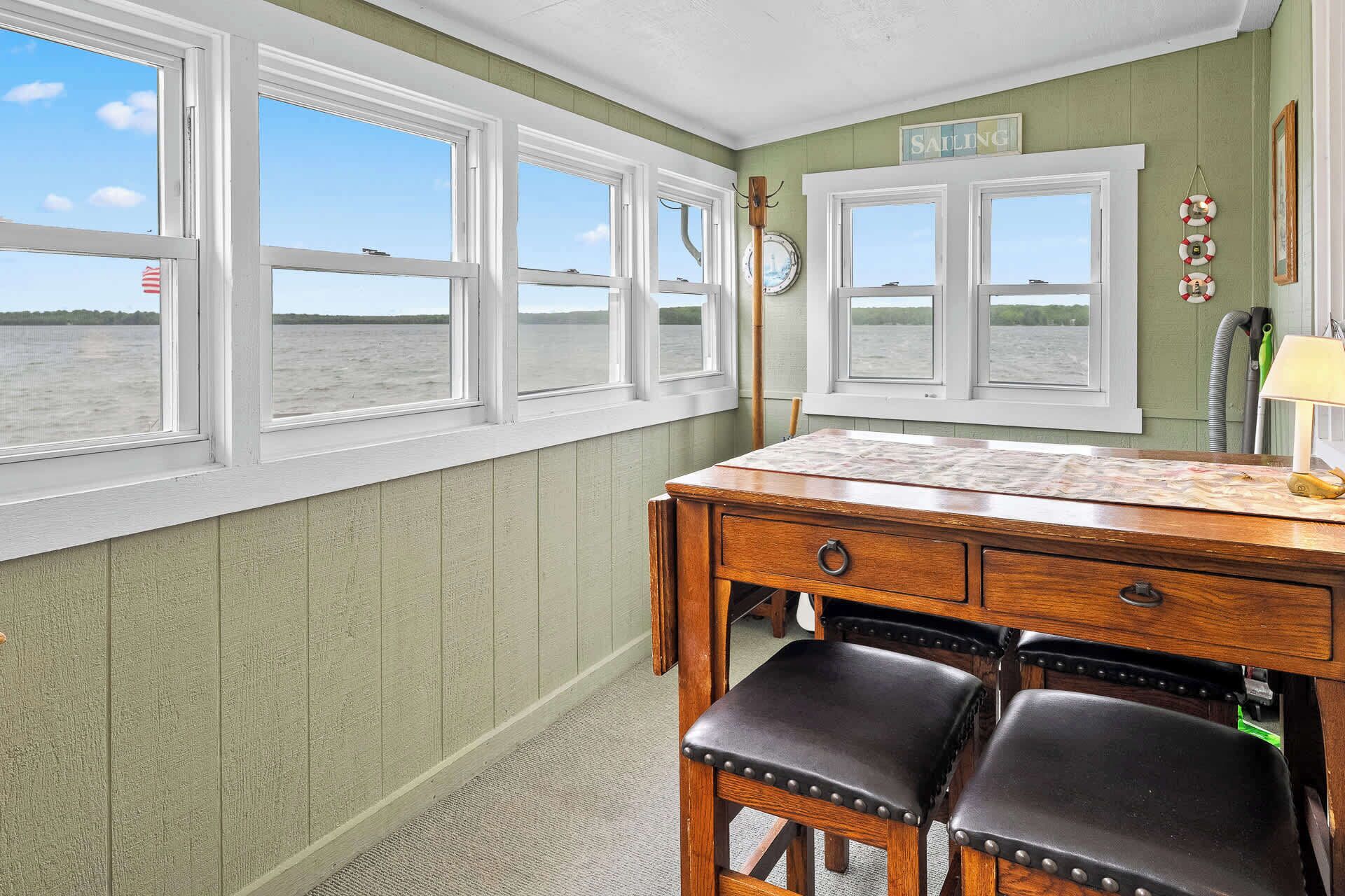 A room with a wooden desk, two leather chairs, and a view of the water through large windows.