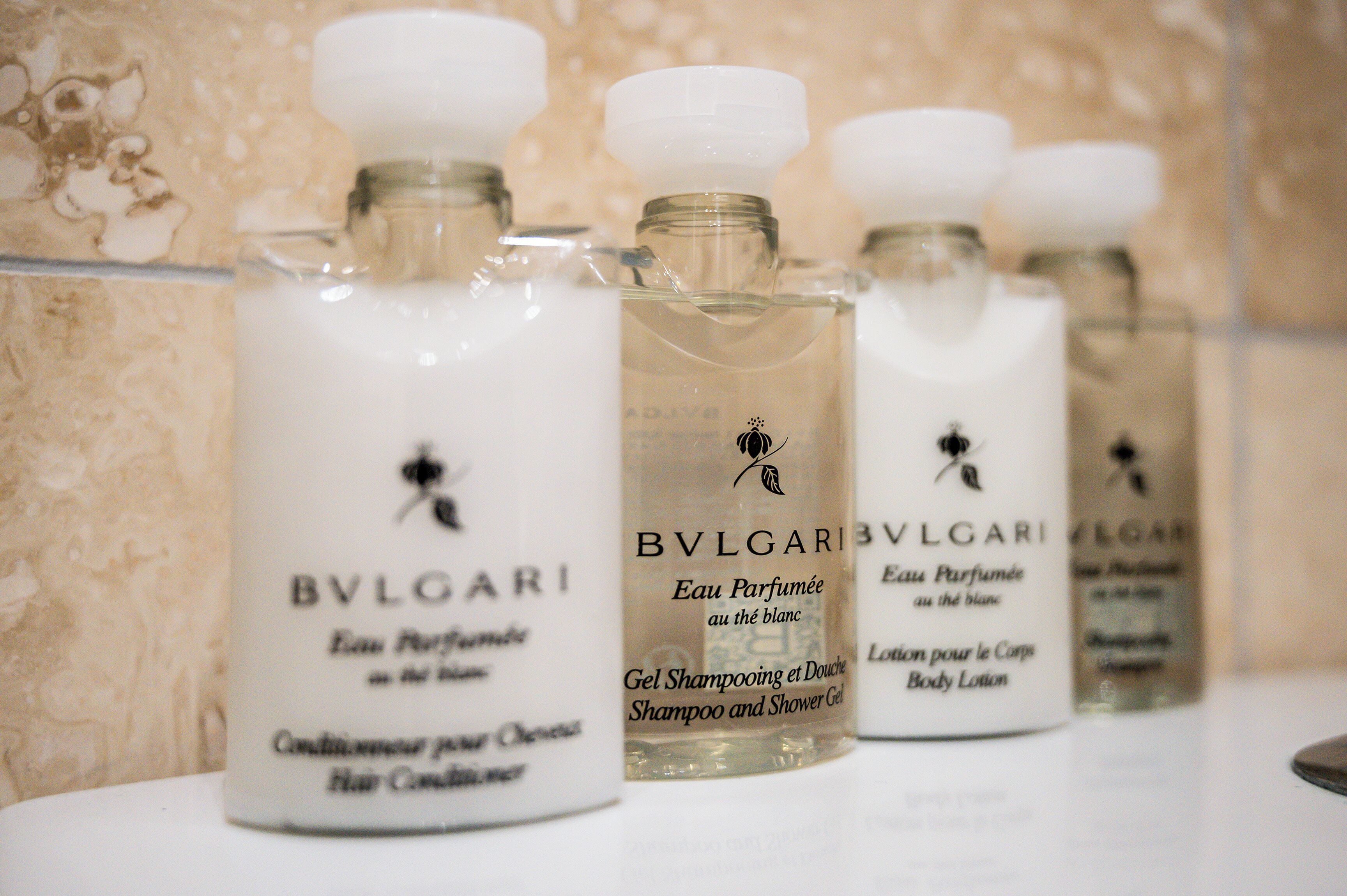 Vários frascos de perfume BVLGARI dispostos em uma superfície branca, com uma parede de azulejos ao fundo.